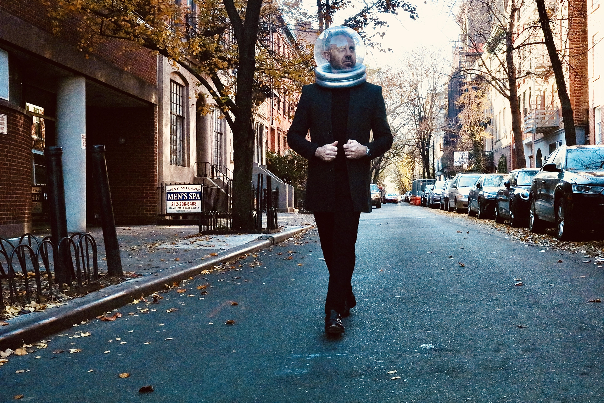 Man in a black coat walking confidently down an urban street wearing a clear astronaut helmet.