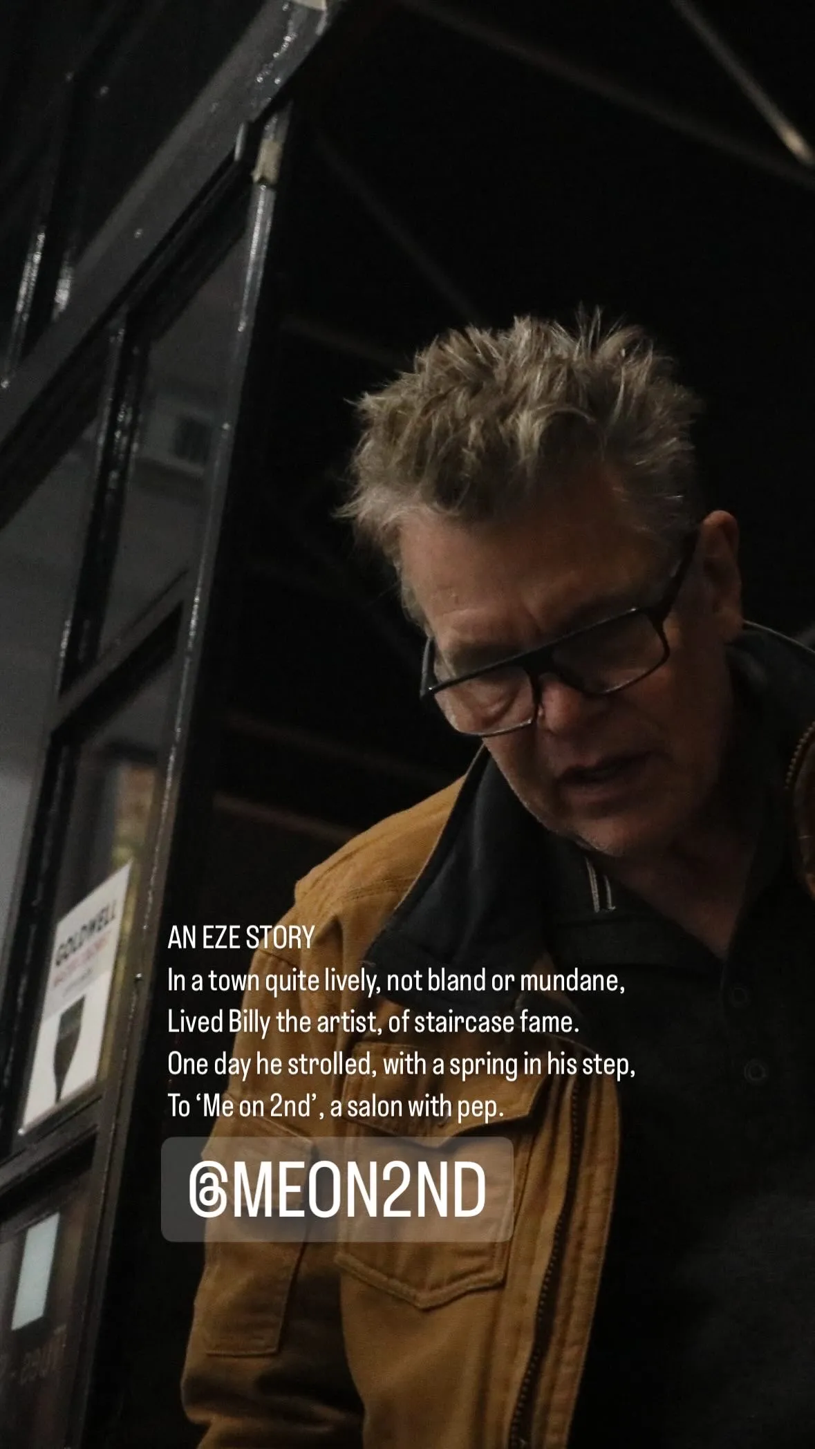 Man wearing glasses and a brown jacket standing near a black door with white text overlay about Billy the artist visiting 'Me on 2nd' salon.