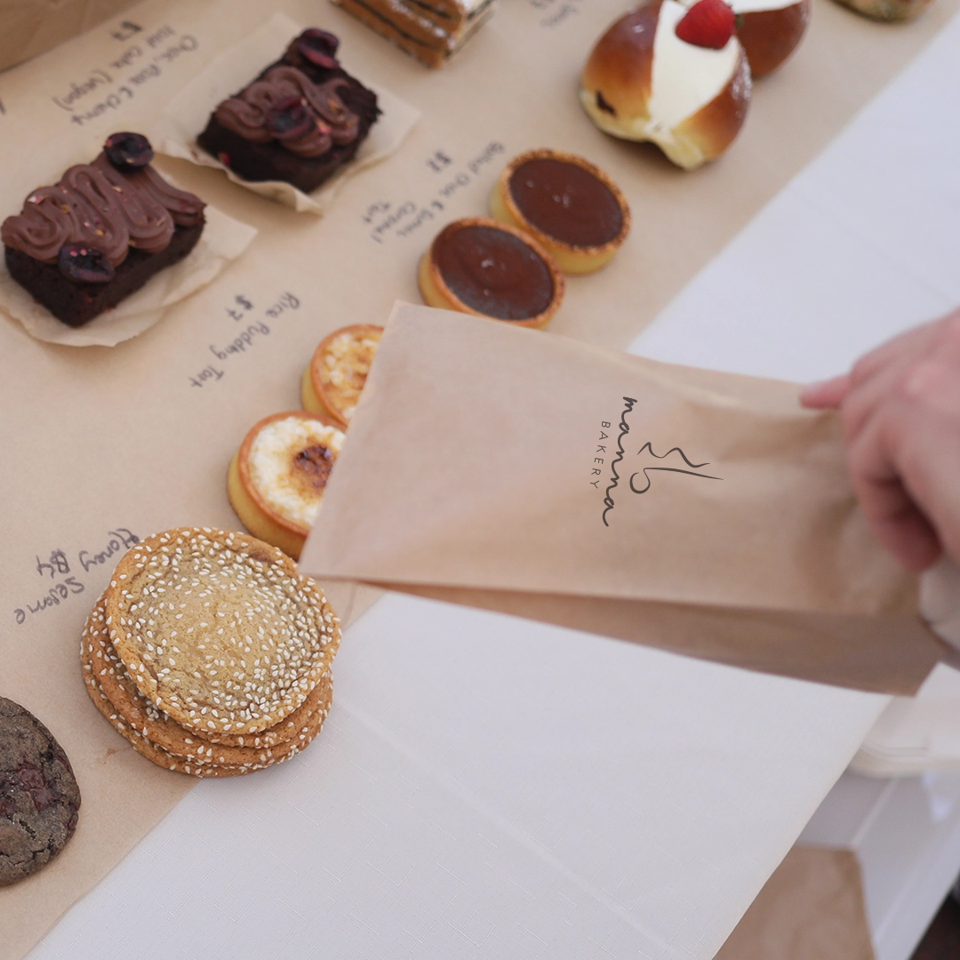 Hand holding a Manna Bakery brown paper bag above assorted cookies and pastries on a table with handwritten labels.