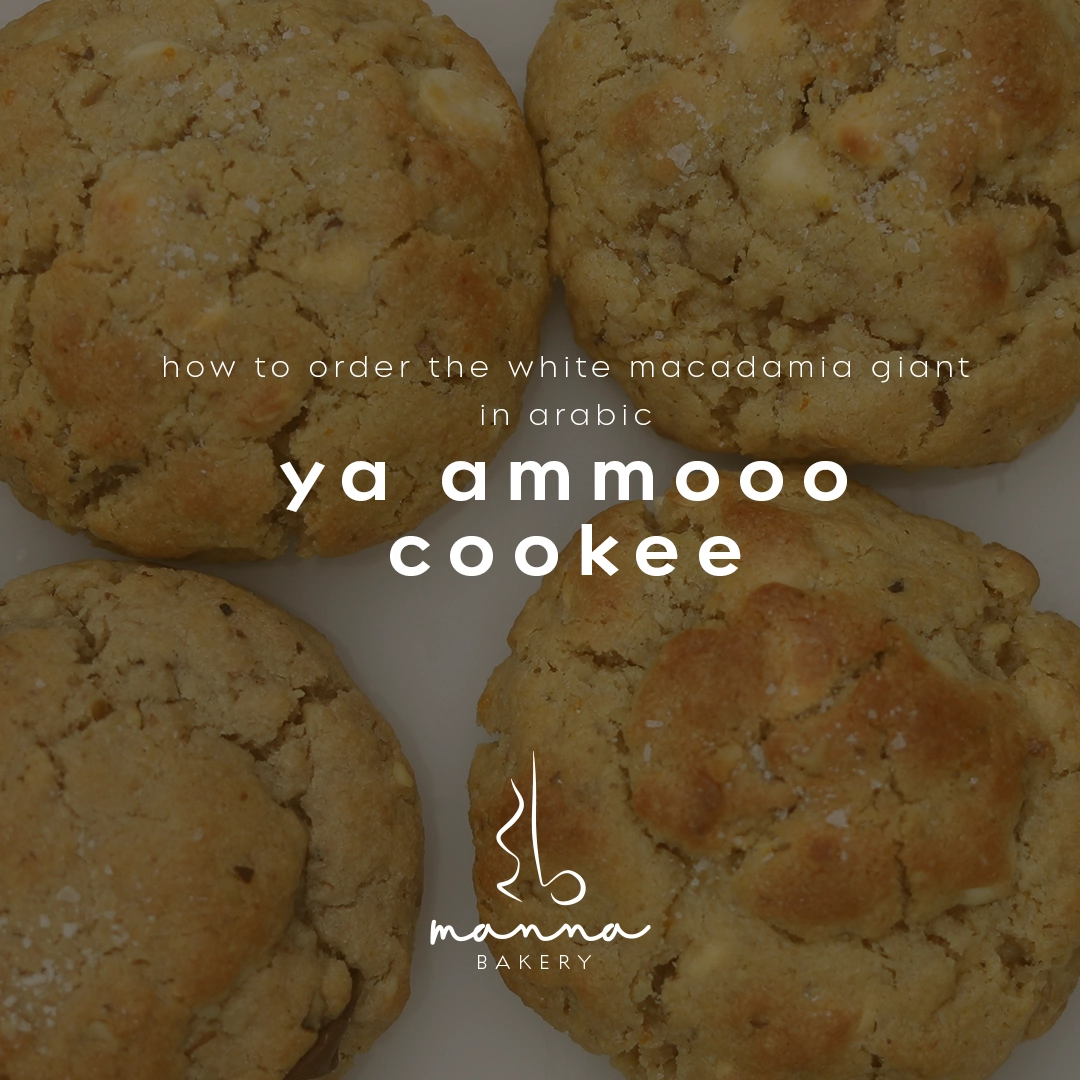 Close-up of four white macadamia giant cookies with text overlay showing the Arabic phrase 'ya ammooo cookee' for ordering, and the Manna Bakery logo.