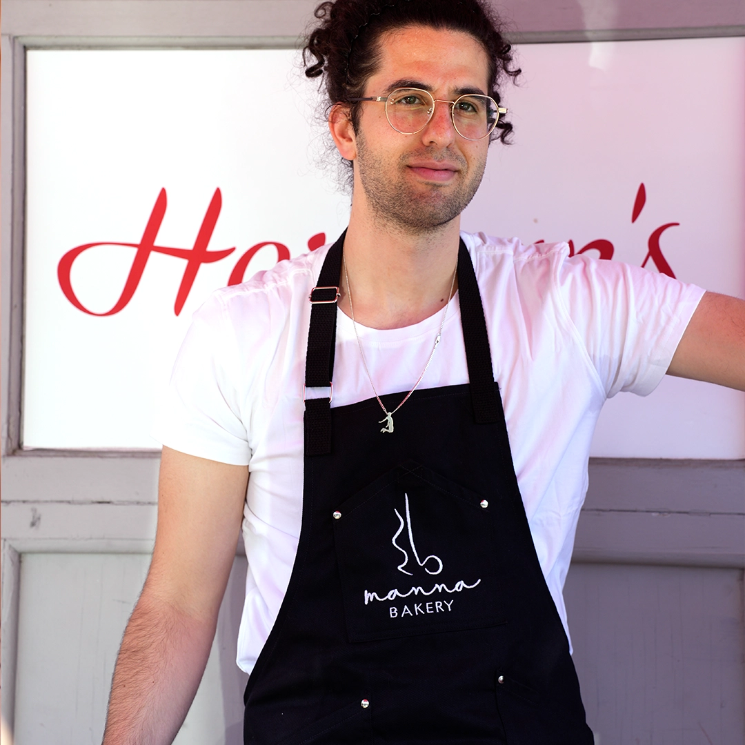 Young man with curly hair and glasses wearing a white T-shirt and black apron that reads 'manna BAKERY', standing in front of a sign with red cursive letters.