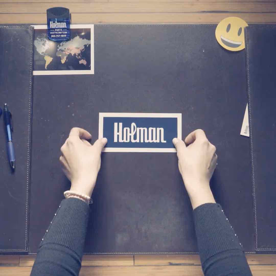 Two hands holding a rectangular card with the word 'Holman' on a dark leather desk mat, a pen on the left, a smiley face paperclip on the top right, and a small world map photo near the top left.