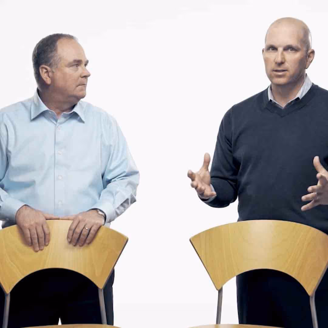 Two men standing behind wooden chairs; the man on the right is speaking and gesturing with his hands, while the man on the left looks at him.
