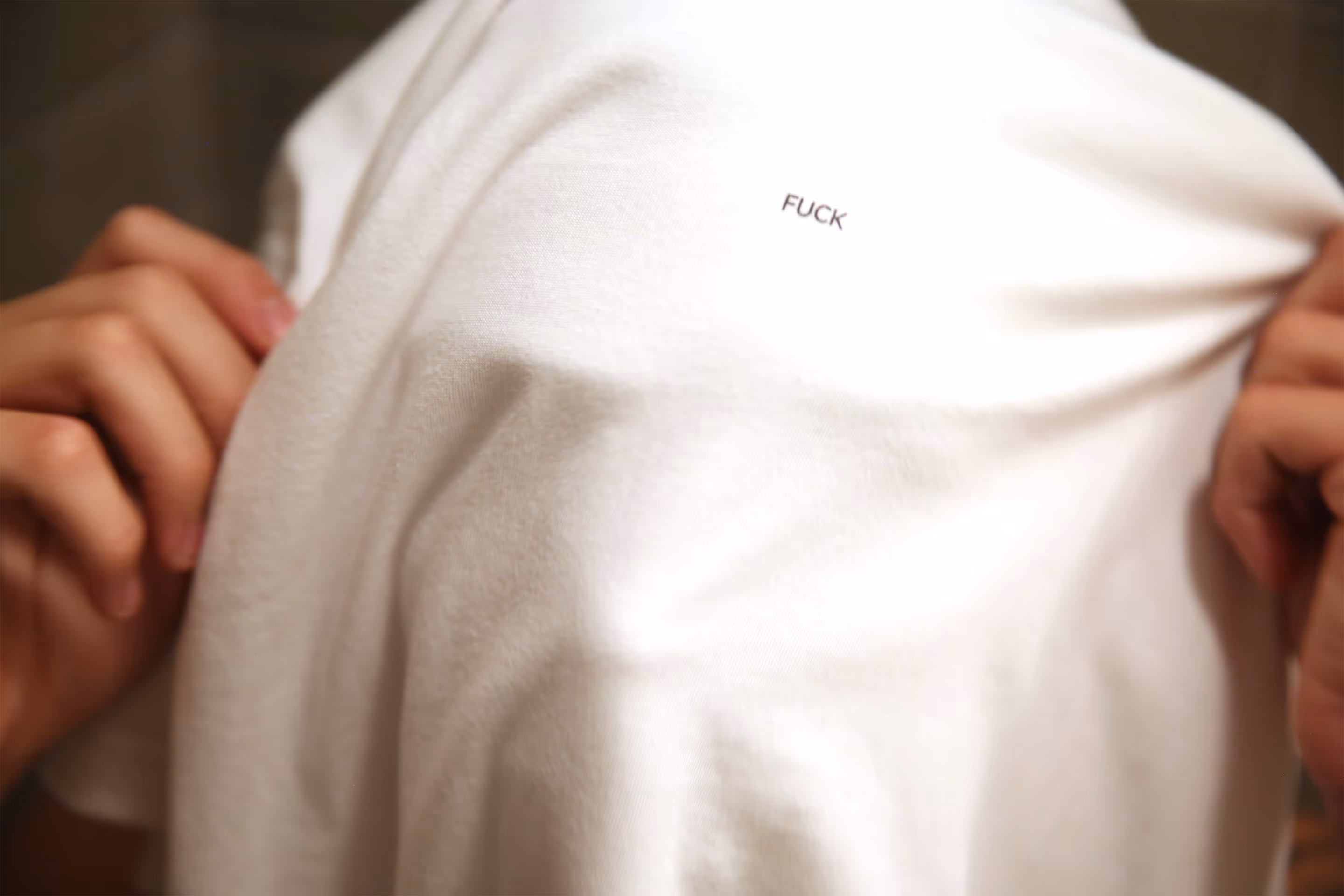 A black and white picture of a person holding a TKINK designer white t-shirt with a small printed "FUCK" over their head.
